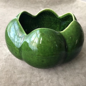 May include: A glossy, green ceramic bowl with a unique, scalloped rim. The bowl's exterior has a smooth, rounded shape, while the interior is a darker shade of green. This decorative bowl is suitable for various uses.