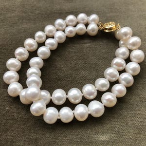 May include: A double-strand pearl bracelet with a gold-colored clasp. The pearls are white and round, strung together to form a classic jewelry piece. The bracelet is displayed on a textured, olive-green surface.