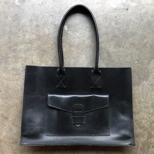 Puede incluir: Un bolso tote de cuero negro con un bolsillo frontal grande y un cierre de hebilla. El bolso tiene dos asas superiores.