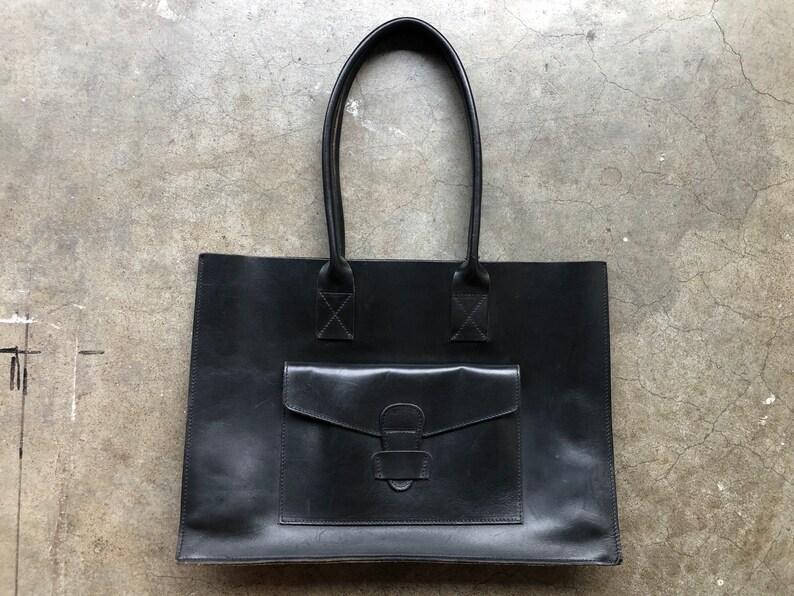 Puede incluir: Un bolso tote de cuero negro con un bolsillo frontal grande y un cierre de hebilla. El bolso tiene dos asas superiores.