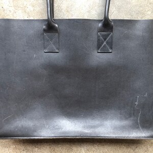 Puede incluir: Un bolso tote de cuero negro con dos asas superiores. El bolso tiene un aspecto desgastado y envejecido.
