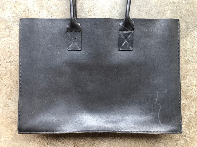 Puede incluir: Un bolso tote de cuero negro con dos asas superiores. El bolso tiene un aspecto desgastado y envejecido.