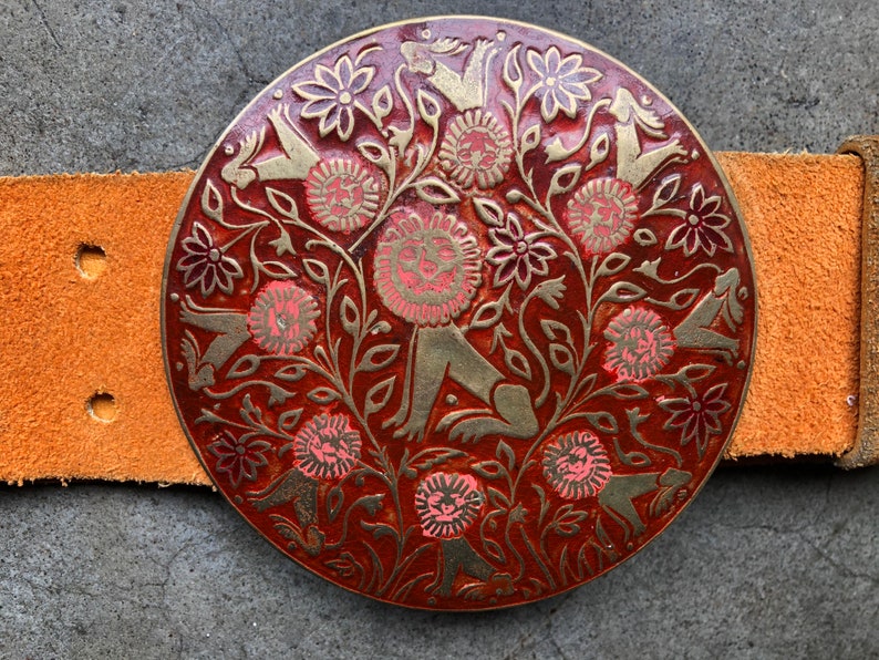 May include: A round, brass belt buckle with a red background and gold-coloured floral and animal designs. The buckle features a repeating pattern of stylised lions and flowers.