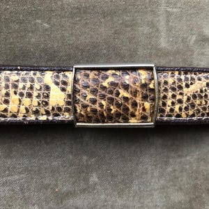 Può includere: Primo piano di una cintura con un motivo a pelle di serpente nei toni del beige, marrone e nero. La cintura ha una fibbia rettangolare color argento. I bordi della cintura sono marrone scuro.