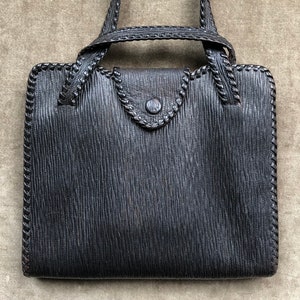 Puede incluir: Un bolso de mano de cuero negro con una correa trenzada y un detalle de puntadas decorativas alrededor de los bordes.