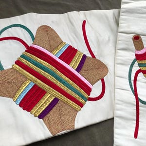 Puede incluir: Dos paneles de tela blanca con diseños bordados. Uno presenta un diseño en forma de cruz con acentos marrones, rojos, dorados, verdes, rosas y morados. El otro panel tiene un diseño en forma de carrete con colores similares y un hilo rojo.