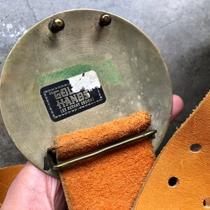May include: A brass belt buckle with a round, circular design. The buckle has a label that reads "Gol Hands Lee Bernay Designs". The buckle is attached to a brown leather belt.