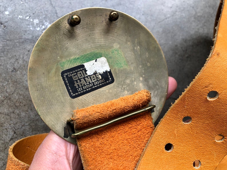 May include: A brass belt buckle with a round, circular design. The buckle has a label that reads "Gol Hands Lee Bernay Designs". The buckle is attached to a brown leather belt.