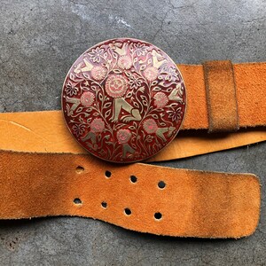May include: A brown leather belt with a round, red and gold buckle featuring a stylised design of animals and flowers.