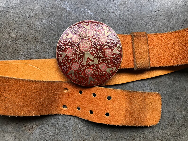 May include: A brown leather belt with a round, red and gold buckle featuring a stylised design of animals and flowers.