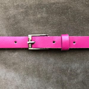 Könnte beinhalten: Ein leuchtend pinkfarbener Gürtel mit einer silberfarbenen Schnalle. Der Gürtel besteht aus glattem Material und hat mehrere Löcher zur Anpassung. Die Schnalle ist rechteckig und hat ein klassisches Design.