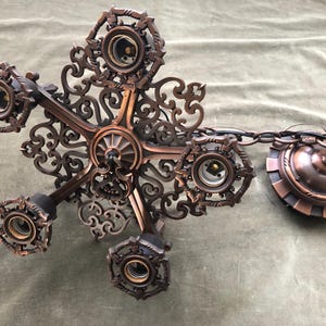 May include: Ornate bronze-colored chandelier with five light sockets. The fixture features intricate scrollwork and a decorative ceiling mount. The light fixture is suspended by a chain.
