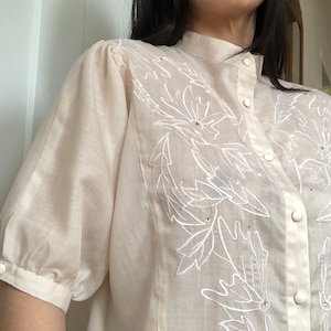 Puede incluir: Una blusa de color crema, transparente, con un diseño de bordado floral blanco. La blusa tiene un cuello mandarín y cierre de botones.