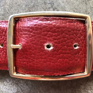 May include: A red leather belt with a gold metal buckle. The buckle is rectangular with rounded corners and has a smooth, polished finish.