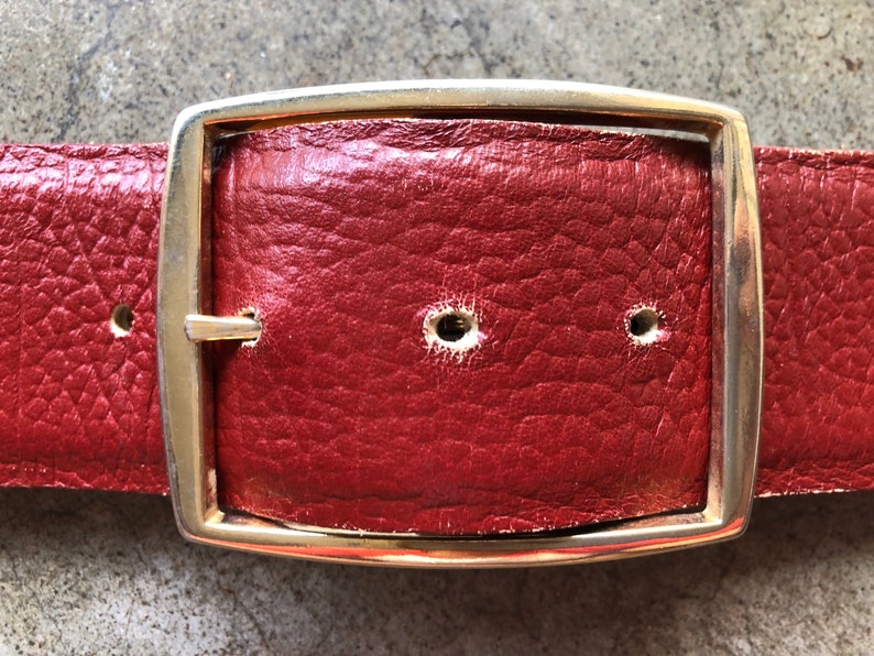 May include: A red leather belt with a gold metal buckle. The buckle is rectangular with rounded corners and has a smooth, polished finish.