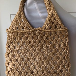 May include: A woven tote bag in a natural tan color. The bag features a macrame-style design with a diamond pattern and a sturdy handle. The interior is lined with a lighter-colored fabric. The bag is designed for carrying items.