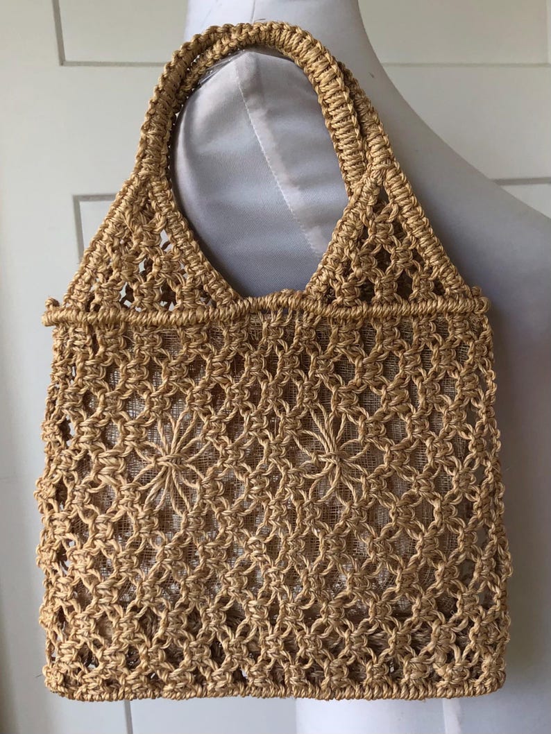 May include: A woven tote bag in a natural tan color. The bag features a macrame-style design with a diamond pattern and a sturdy handle. The interior is lined with a lighter-colored fabric. The bag is designed for carrying items.