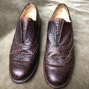 May include: A pair of brown leather shoes with a brogue design. The shoes have a textured, alligator-like pattern.