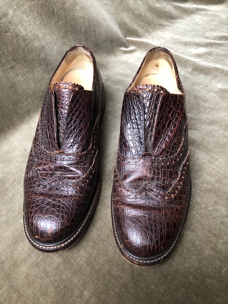 May include: A pair of brown leather shoes with a brogue design. The shoes have a textured, alligator-like pattern.
