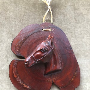 May include: A wooden horse head sculpture mounted on a brown wood plaque with a natural twine hanger.