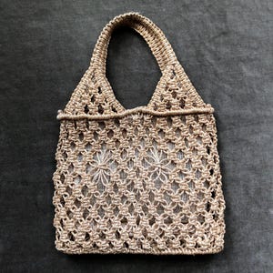 May include: A beige macrame tote bag with a woven design. The bag has a rectangular body with a handle and is made of a natural fiber. The bag is empty and sits on a dark gray surface.