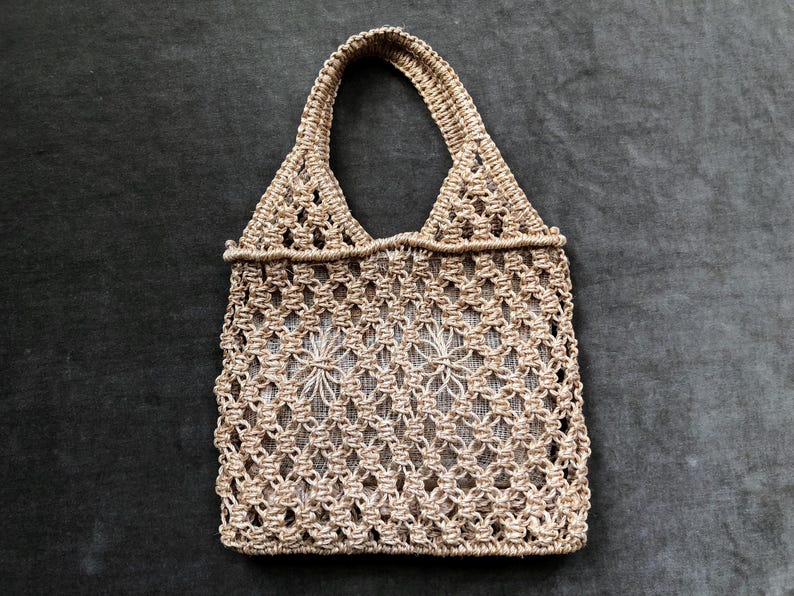 May include: A beige macrame tote bag with a woven design. The bag has a rectangular body with a handle and is made of a natural fiber. The bag is empty and sits on a dark gray surface.