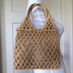 Puede incluir: Un bolso tote de macramé beige con un diseño floral tejido. El bolso tiene forma rectangular con un asa resistente y un patrón tejido. El bolso está hecho de un material de fibra natural.