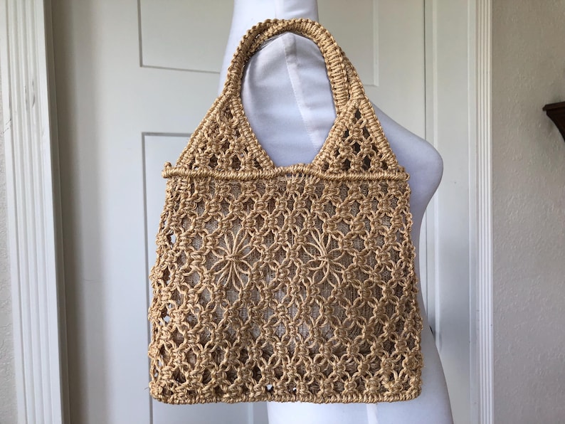 May include: A beige macrame tote bag with a woven floral design. The bag has a rectangular shape with a sturdy handle and a woven pattern. The bag is made of a natural fiber material.