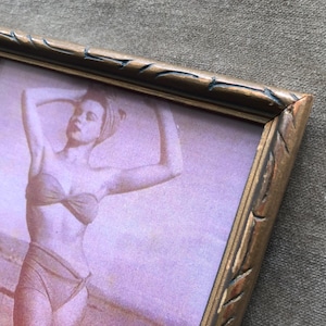 May include: A rectangular picture frame with a distressed bronze and black finish featuring carved diagonal notches along the edges. The frame contains a sepia-toned photograph of a person in a two-piece swimsuit posing on a beach.