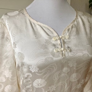 May include: A white silk blouse with a floral pattern and a mandarin collar. The blouse has a button closure at the neck.