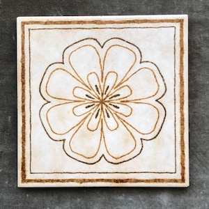 May include: Square ceramic tile with a floral design. The tile features a cream-colored background with a brown floral pattern in the center and a brown border. The floral design is symmetrical and detailed.