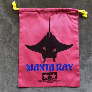 Puede incluir: Una bolsa de cordón rosa con una silueta de manta raya negra y el texto "MANTA RAY" en azul. La bolsa también presenta el logotipo de Tamiya Plastic Model Co. y el texto "Hecho en Japón".