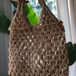 May include: A woven, open-weave tote bag in a natural tan color. The bag features a sturdy handle and a decorative, open pattern. The bag is designed with a rectangular shape and is suitable for carrying various items.