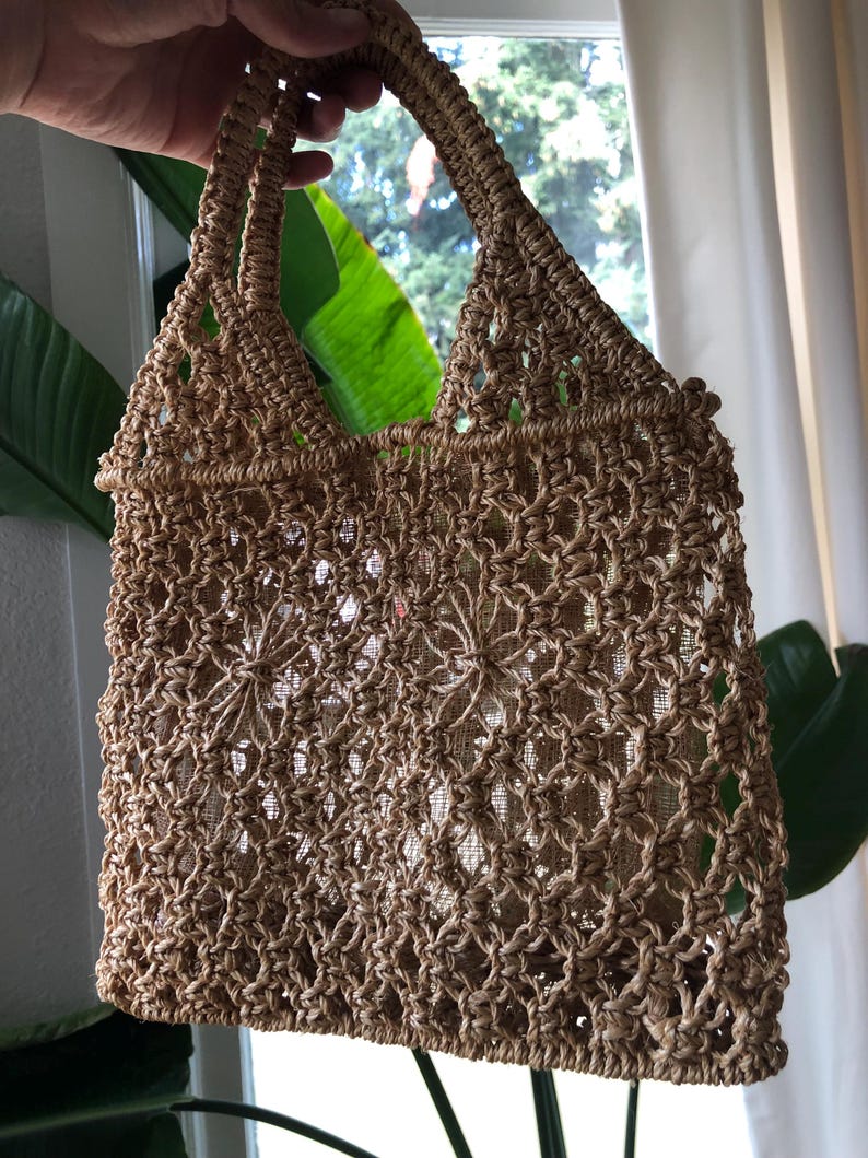 May include: A woven, open-weave tote bag in a natural tan color. The bag features a sturdy handle and a decorative, open pattern. The bag is designed with a rectangular shape and is suitable for carrying various items.
