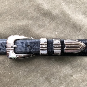 May include: A close-up of a dark blue belt with a silver buckle and accents. The belt has a textured, reptile-like pattern. The buckle and belt loops show signs of wear, adding a vintage aesthetic.