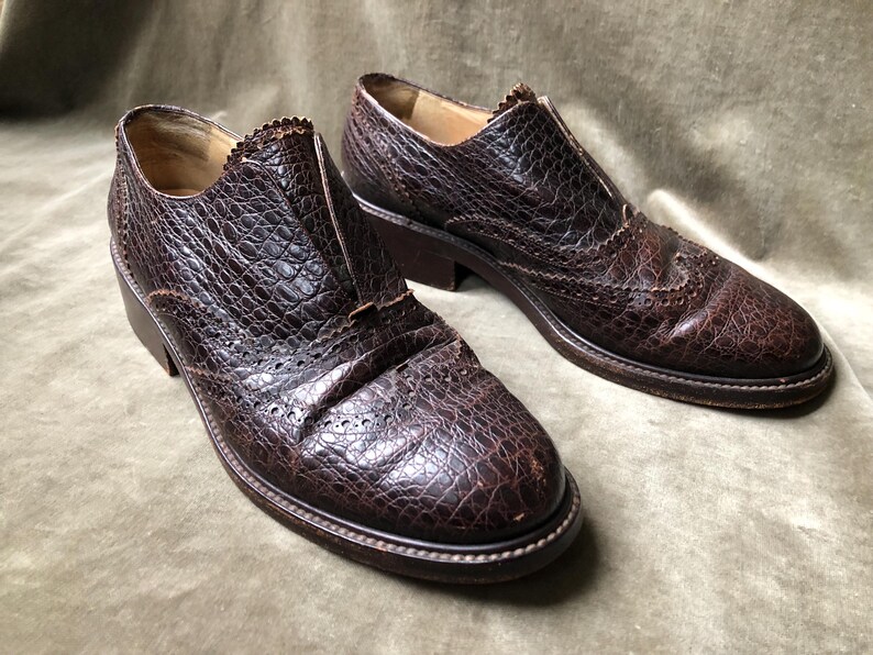May include: A pair of brown leather shoes with a textured, crocodile-like pattern. The shoes have a lace-up front and a wingtip design.