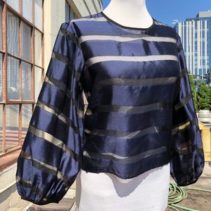 May include: A navy blue blouse with horizontal sheer black and silver stripes. The long sleeves have a balloon shape at the wrist. The blouse has a round neckline and is made of a shiny, lightweight fabric.