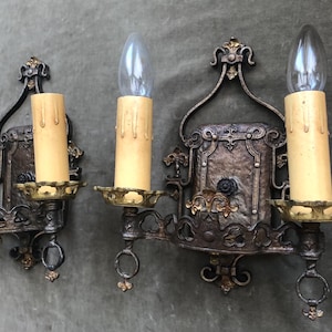 May include: Ornate, antique-style wall sconces. Each sconce features a candle-shaped light bulb, a faux candle, and decorative metalwork in shades of bronze and gold. The sconces are mounted on a wall.
