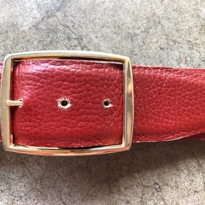 May include: A red leather belt with a gold metal buckle. The buckle is rectangular with rounded corners.