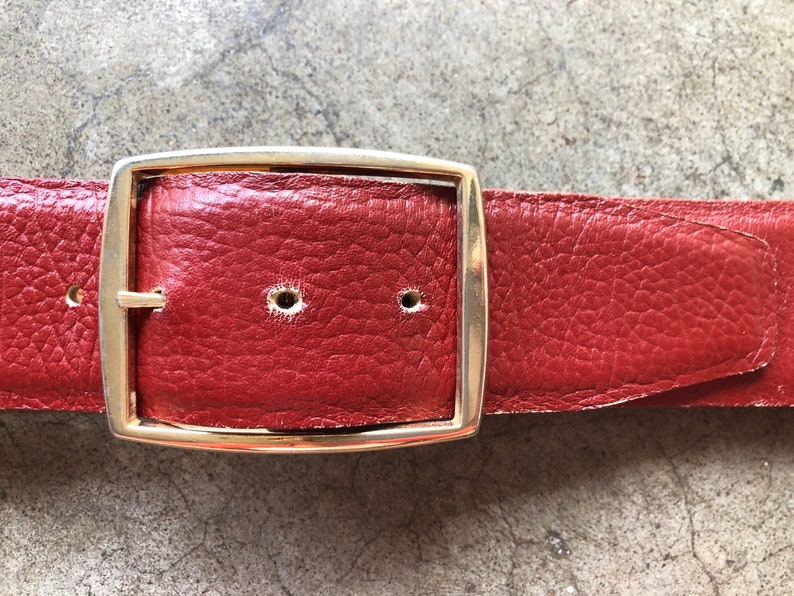 May include: A red leather belt with a gold metal buckle. The buckle is rectangular with rounded corners.