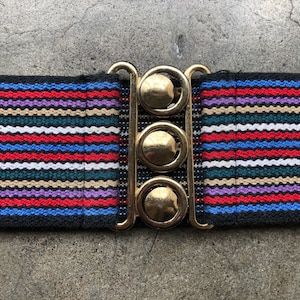 Puede incluir: Un cinturón elástico negro con un patrón de rayas coloridas en rojo, azul, blanco, morado y verde. El cinturón tiene una hebilla de metal dorado con tres piezas redondas decorativas.