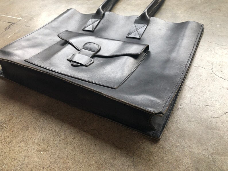 Puede incluir: Un bolso tote de cuero negro con un bolsillo frontal grande y dos asas superiores.
