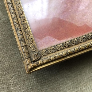 May include: A rectangular antique picture frame with a gold finish and ornate floral detailing. The frame surrounds a pink and white image. The frame is made of metal and has a vintage aesthetic.