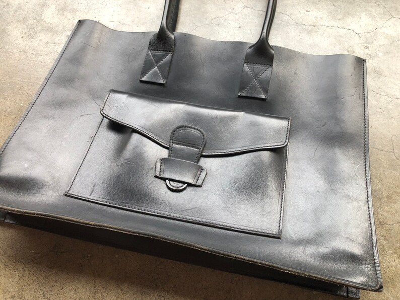 Puede incluir: Un bolso tote de cuero negro con un bolsillo frontal grande y dos asas superiores.