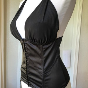 Pode incluir: Corpete de cetim preto com decote halter e ferragens douradas. O corpete tem uma estrutura com varas e uma borda de renda.