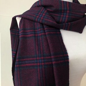 May include: A long rectangular scarf featuring a dark navy blue, deep red, and forest green plaid pattern. The fabric displays a woven texture with a classic grid design, draped around a white mannequin neck, showcasing the intricate color intersections.