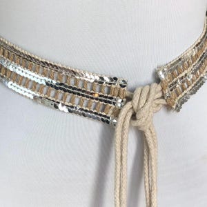 May include: A beige rope belt featuring three horizontal rows of shiny silver sequins woven into the fabric. The belt is secured with a simple knot, and two long, loose rope ends hang down from the center, creating a textured, layered appearance.