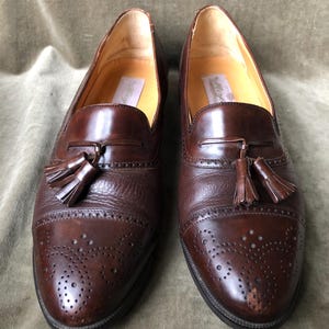 May include: A pair of brown leather tassel loafers with a wingtip design and a perforated pattern. The loafers have a brown leather sole and a tassel detail on the front.