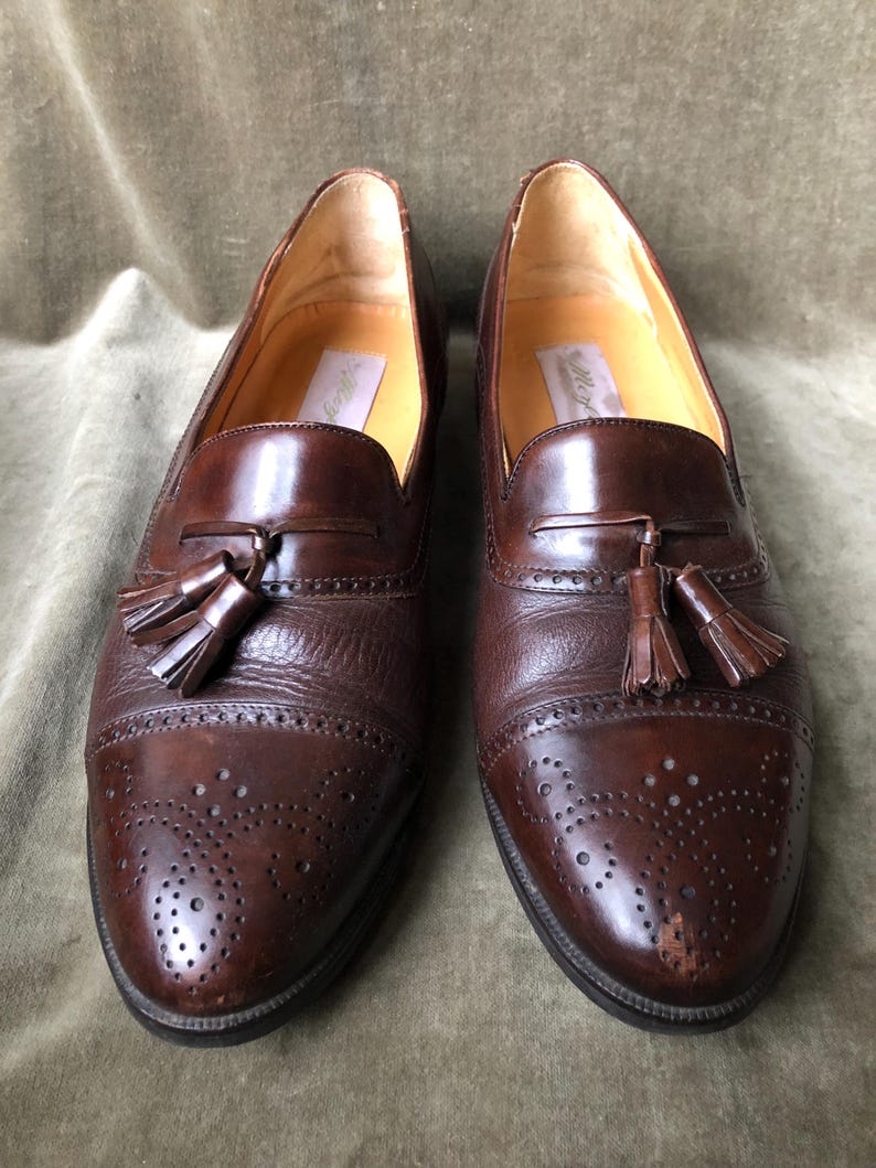 May include: A pair of brown leather tassel loafers with a wingtip design and a perforated pattern. The loafers have a brown leather sole and a tassel detail on the front.