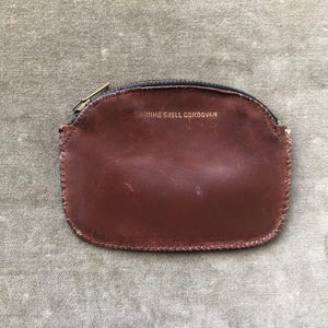 May include: A dark brown leather coin purse with a semi-circular shape and a top zipper closure. The front surface features the embossed text GENUINE SHELL CORDOVAN in gold lettering. The edges are stitched with a visible thread pattern.